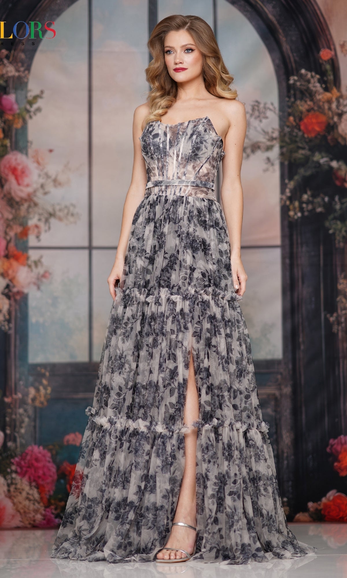 A woman stands before a floral backdrop in the Strapless Long Print Prom Dress: Colors Dress 3810, featuring a strapless design, front slit, and paired with silver strappy heels.